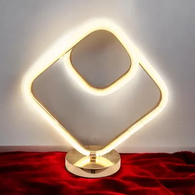 أباجورة LED ذهبية بتصميم هندسي عصري، حيث تتكون من شكلين مربعين متداخلين و بقاعدة ذهبية لامعة ويبدو مضاء باللون الأبيض الدافئ