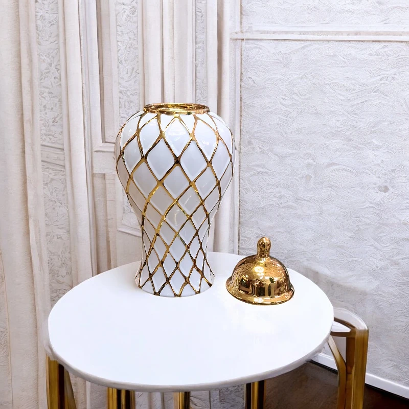 فازة زنجبيل سيراميك ذهبية بتصميم مربعات زخرفية - الحجم الوسط، موديل 29143 فازة زنجبيل زخرفية تتميز بنقوش ذهبية هندسية وغطاء دائري ذهبي يظهر بجانب الفازة على الترابيزة.