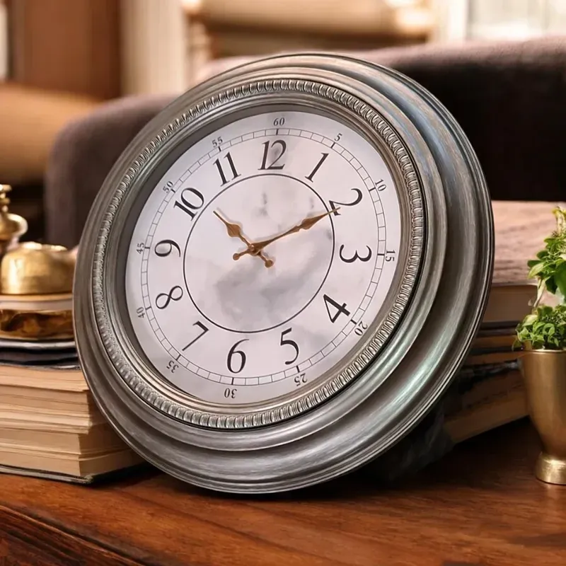 ساعة حائط دائرية، إطار فضي، ميناء أبيض عاجي، أرقام عربية سوداء، عقارب ذهبية.
