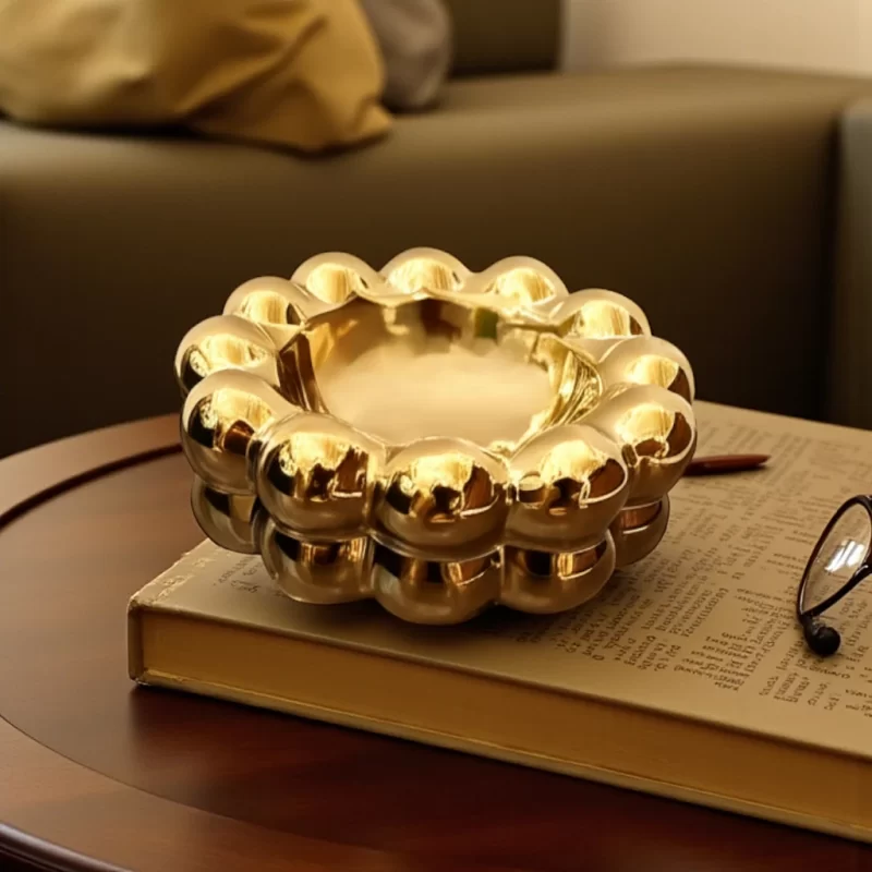 إخوان سرور الإضاءة الحديثة والديكور صورة لطفاية بورسلين ذهبية فاخرة بتصميم فقاعات، موضوعة على كتاب فوق طاولة خشبية.