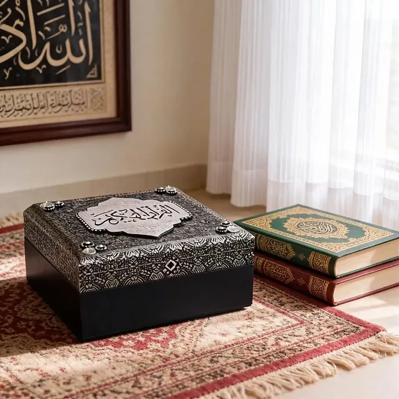 صندوق حفظ مصحف إسلامي فاخر بنقوش فضية كريستالية وغطاء أسود وجسم أزرق داكن مغلق وموضوع بجانب مصاحف.